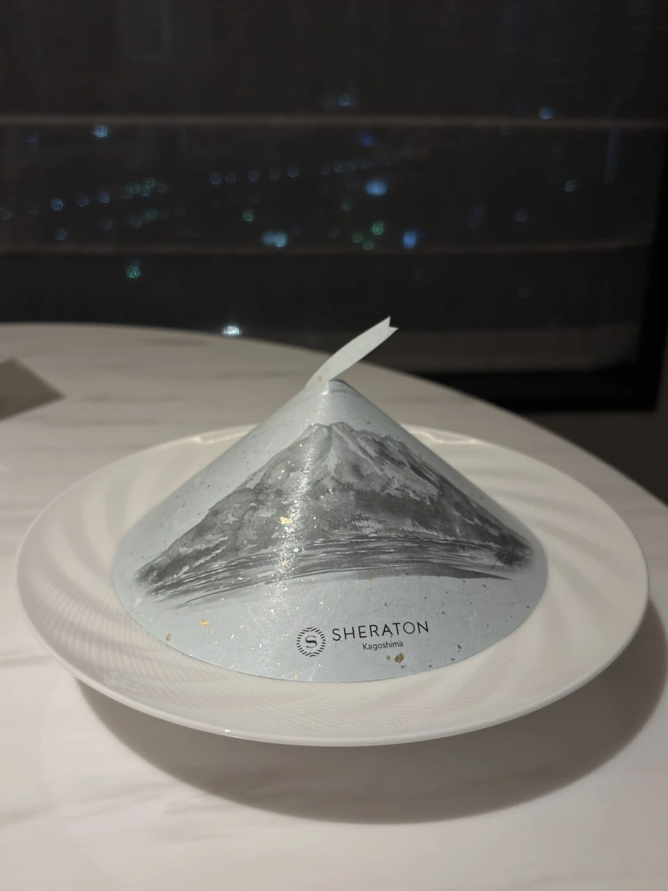 桜島の絵が描かれた円すい形の紙のカバーをかぶせたお皿。SHERATON Kagoshimaの文字