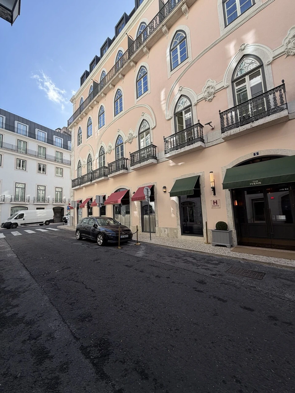 晴れた日のRua Capelo。淡いピンクの建物にジ・アイヴェンスの緑と赤のひさしが並び、細い石畳の道が続く