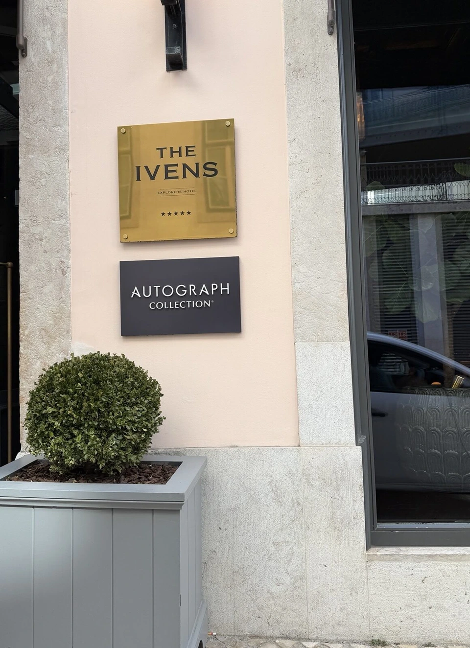 ジ・アイヴェンスの入口の壁。THE IVENS EXPLORERS HOTELの金のプレートとAUTOGRAPH COLLECTIONの黒い看板、丸く刈った植木
