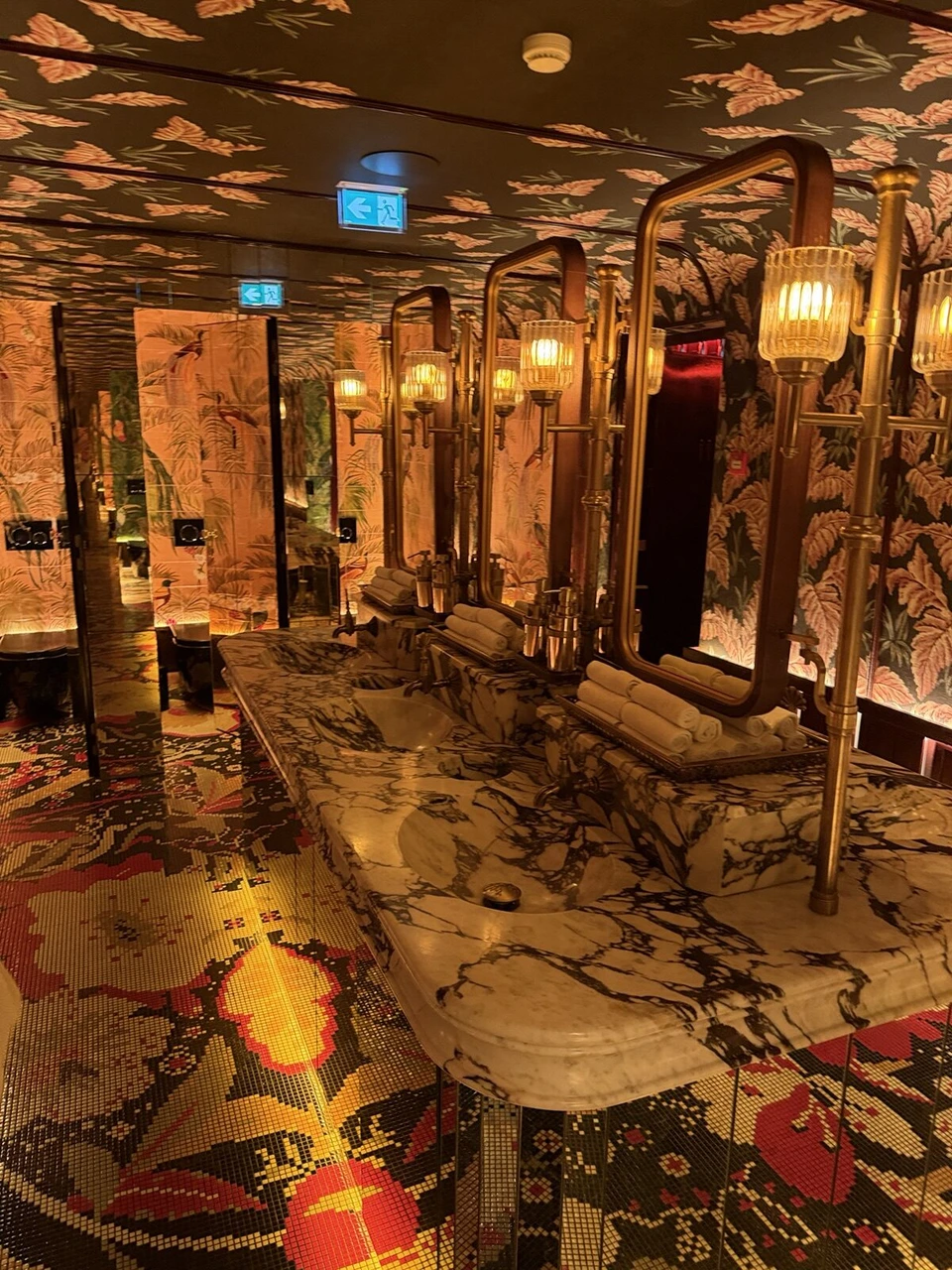 ジ・アイヴェンスの館内の化粧室。熱帯の植物柄の壁紙と天井、大理石の洗面台、金色の照明と鏡、模様のモザイクの床