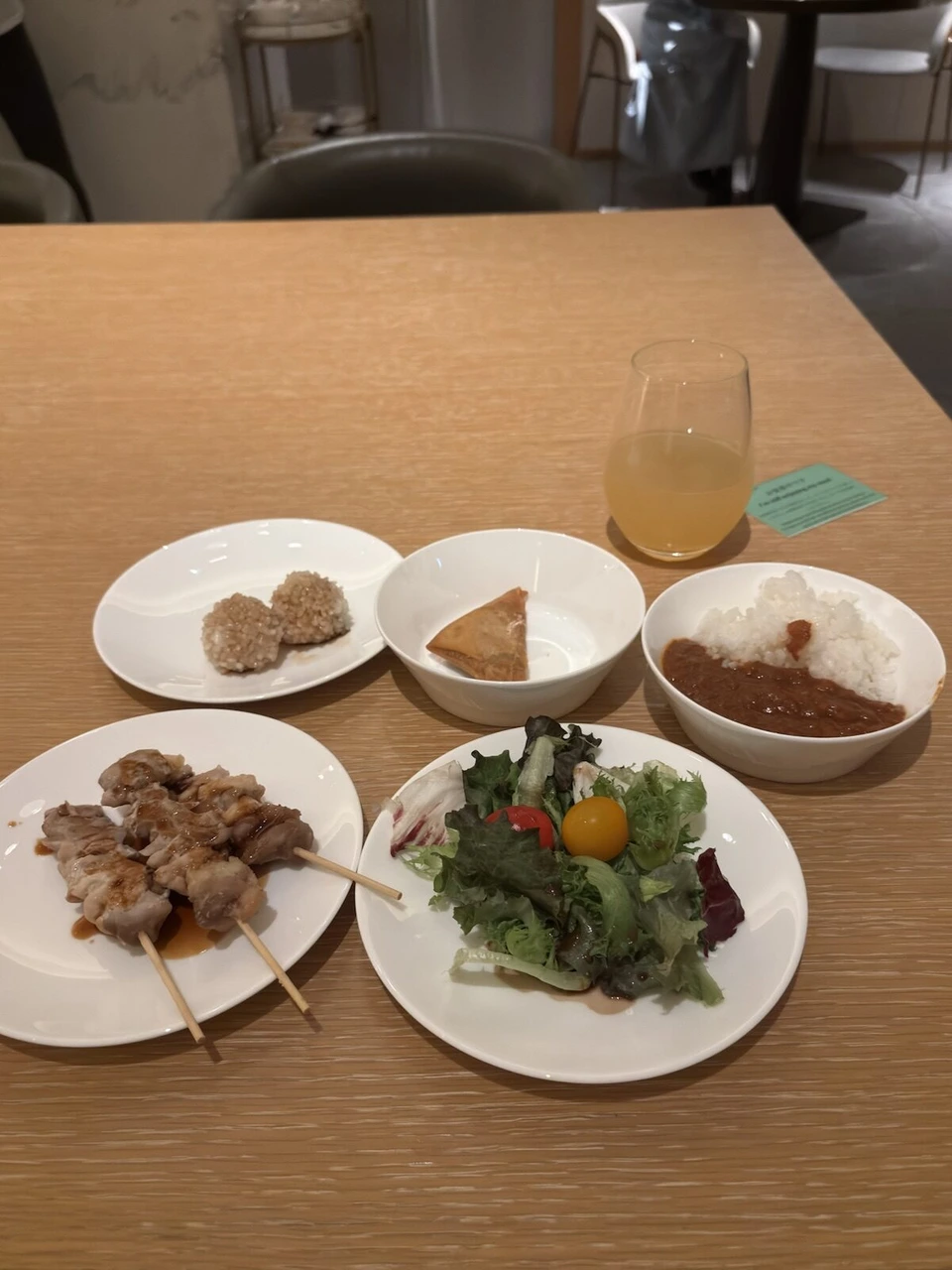 クラブラウンジのテーブルに並んだ焼き鳥、サモサ、カレーライス、サラダ、黒米のおにぎりのような一品とジュース