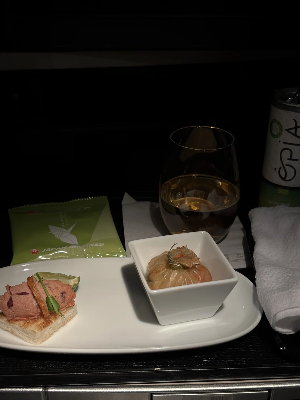 JL724ビジネスクラスの最初のサービス。肉のパテをのせたトースト、小さな器の前菜、グラスの飲み物とおつまみの袋