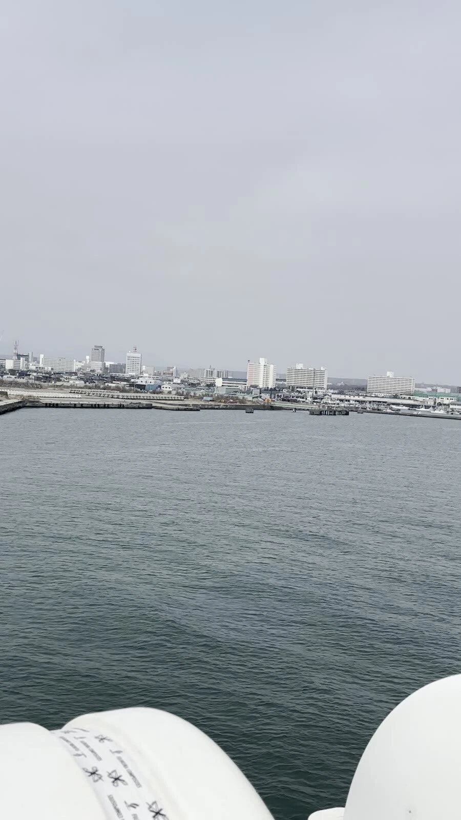 フェリーのデッキから見た苫小牧の港と街並み。曇り空と灰色の海、手前に白い救命いかだのコンテナ