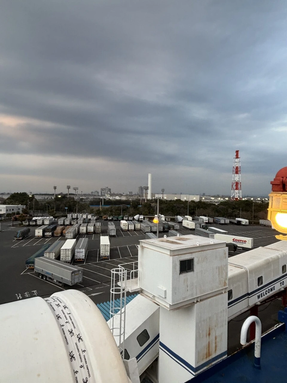 出港前のデッキから見下ろした名古屋港の岸壁。トレーラーが並び、奥に紅白の鉄塔