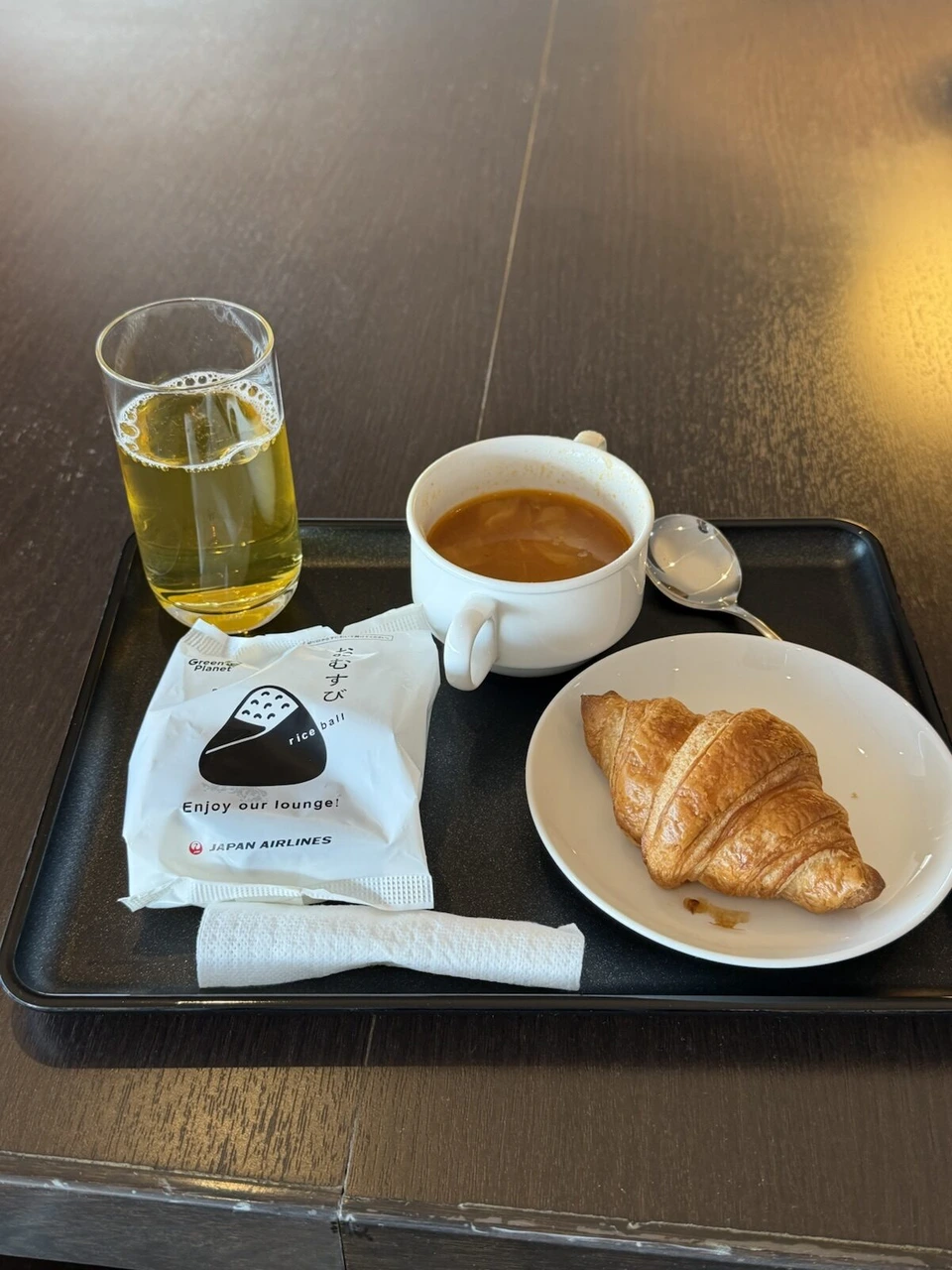 羽田のJALラウンジのトレー。緑茶、スープ、Enjoy our lounge!と書かれたおむすびの包み、クロワッサン