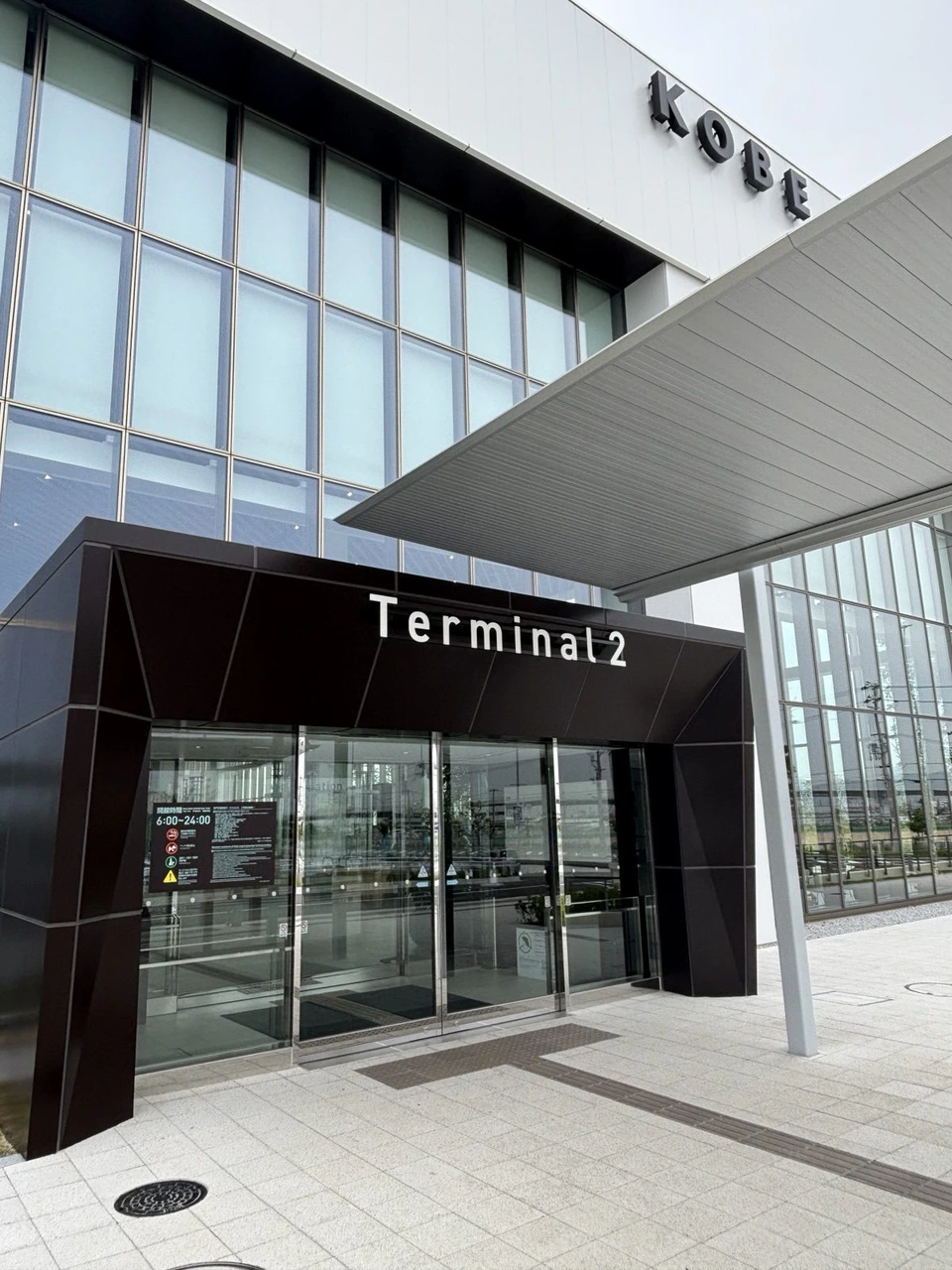 神戸空港第2ターミナルの入口。黒い外壁にTerminal 2の白い文字、ガラス張りの建物の上にKOBEの文字。扉の横に開館時間の案内