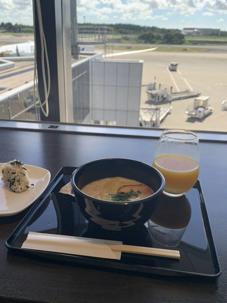 成田空港第1ターミナルのラウンジの窓際席。麺のどんぶり、おにぎり、オレンジジュースと駐機場