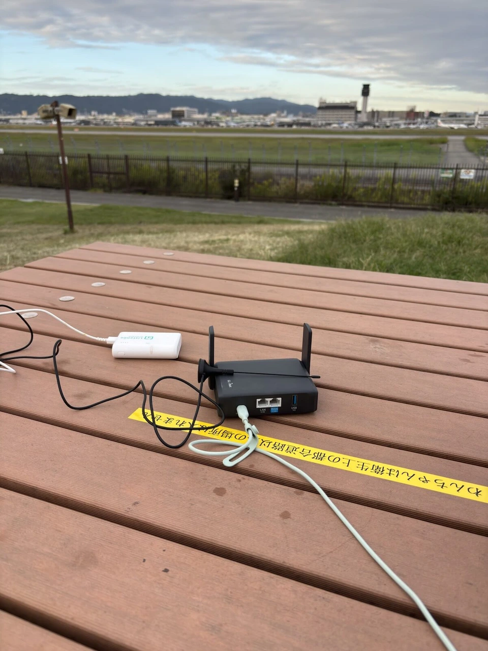 公園の木のテーブルに置いた黒い小型Wi-Fiルーター。アンテナを2本立てていて、奥に白いモバイルバッテリー、その先に伊丹空港の滑走路と管制塔