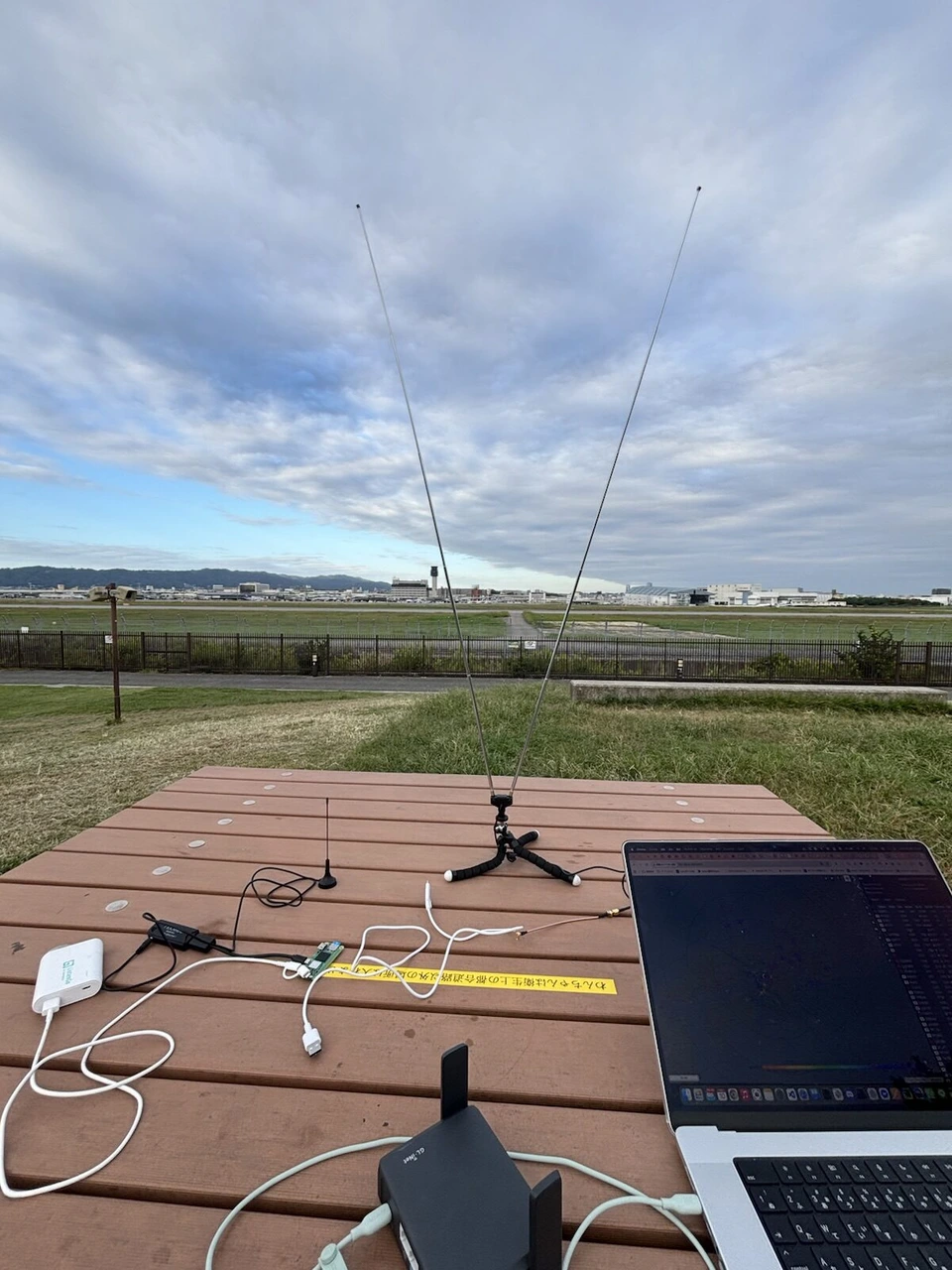 伊丹スカイパークのテーブルに広げた機材。V字のアンテナ、小さな基板、白いモバイルバッテリー、ノートPC、手前に黒いSlate 7