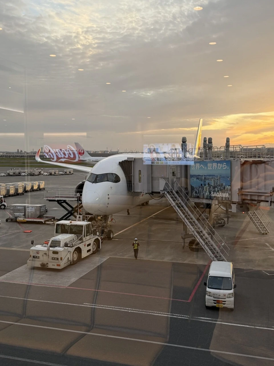 羽田空港のガラス越しに見た夕方のスポット。ボーディングブリッジにつながった白い旅客機と、オレンジ色に染まる雲
