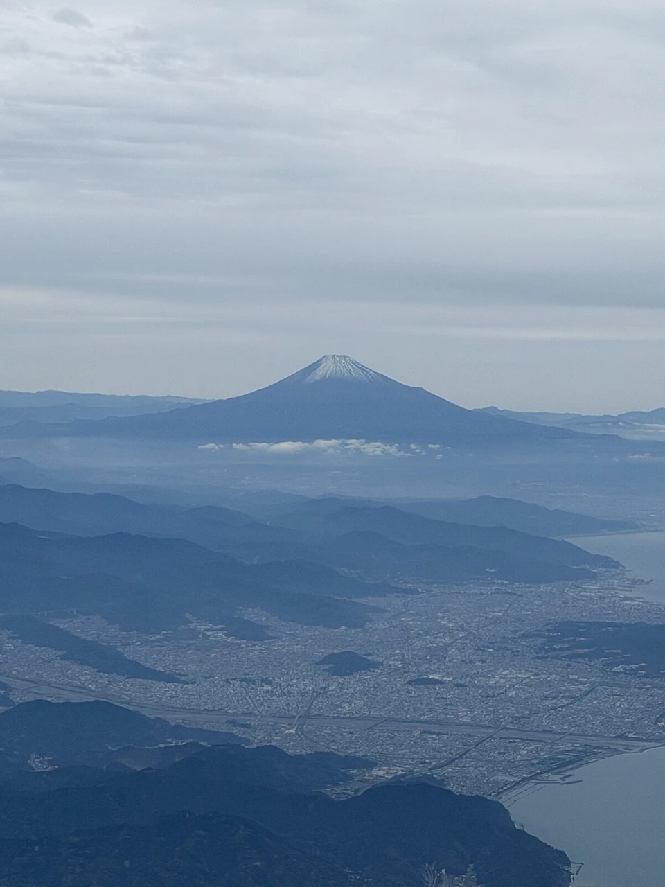 飛行機の窓から見た、雲の上に頭を出す雪をかぶった富士山と、手前の街と海岸線