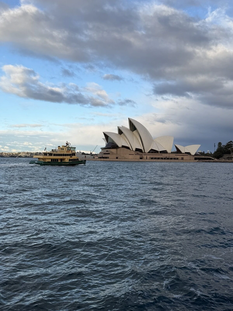 曇り空の下のシドニー・オペラハウスと、手前の海を走る黄色と緑のフェリー