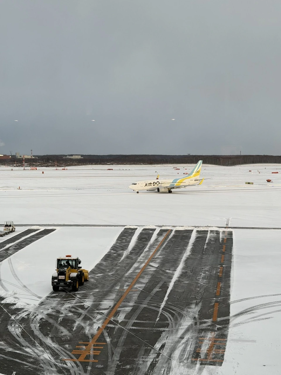 雪で真っ白な新千歳空港の駐機場を走るAIRDOの飛行機と、手前の除雪車