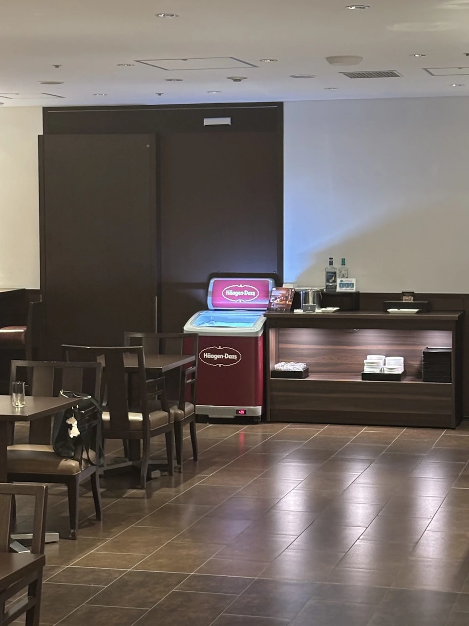 羽田空港第1ターミナルのJALのラウンジ。木目の壁の前にハーゲンダッツのアイスケースと飲み物の棚、手前にテーブル席