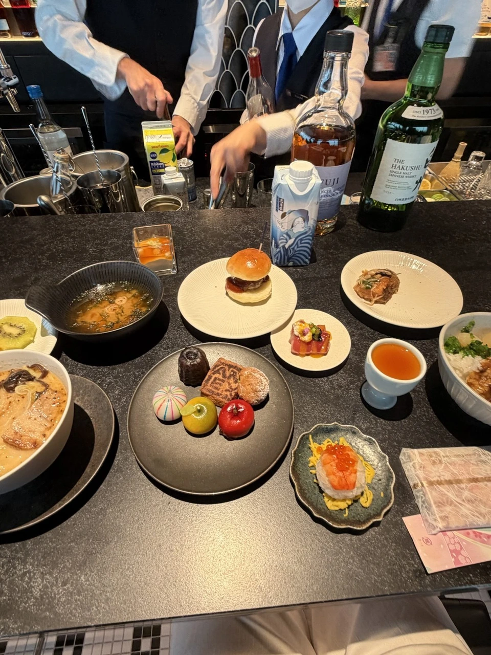 羽田空港第3ターミナルのJALのラウンジのバーカウンター。ラーメンや小さなバーガー、和菓子の皿とウイスキーの瓶