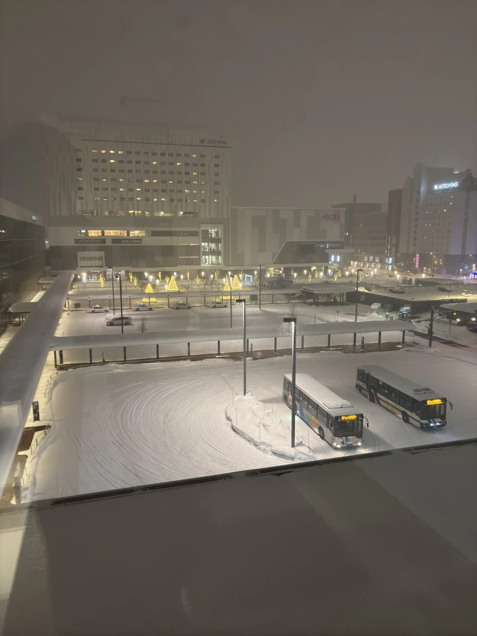 ホテルの窓から見下ろした夜の旭川駅前。雪の積もったバスターミナルに2台のバス、奥にイオンモールと金色のツリーのイルミネーション