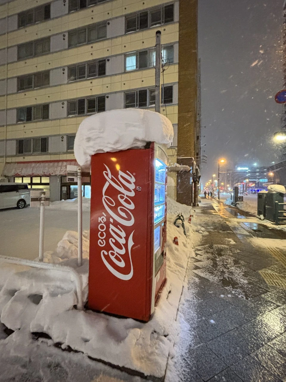 歩道の角に立つコカ・コーラの赤い自動販売機。上に分厚い雪が帽子のように積もっている