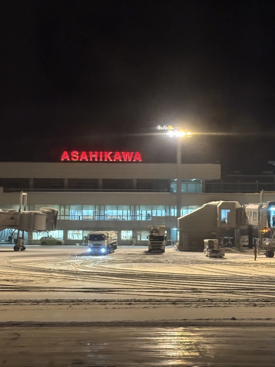 着陸後の機内から見た夜の旭川空港。ターミナルの屋上に赤く光るASAHIKAWAの文字、雪の積もった駐機場とボーディングブリッジ