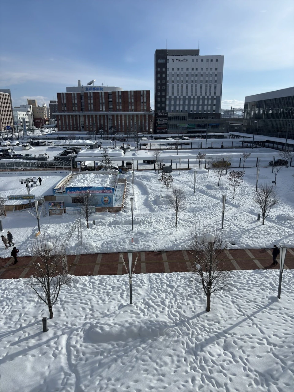 朝のホテルの窓から見た晴れた旭川駅前。青空の下、雪で真っ白な広場と木々、奥に赤れんがの建物とビル