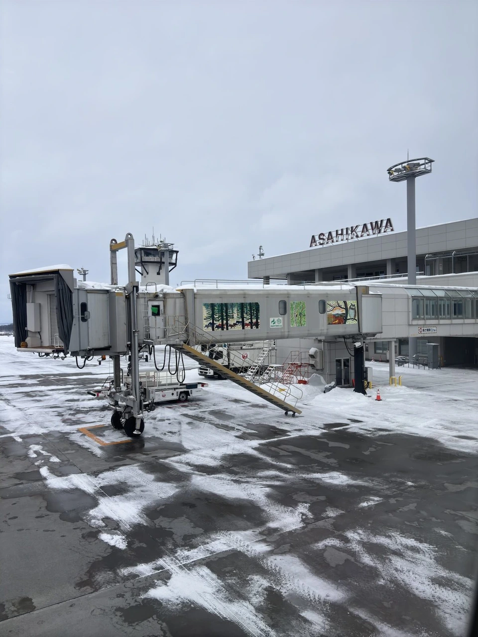 旭川空港の搭乗口から見た、雪の残る駐機場とボーディングブリッジ。奥にASAHIKAWAの文字のターミナルと管制塔