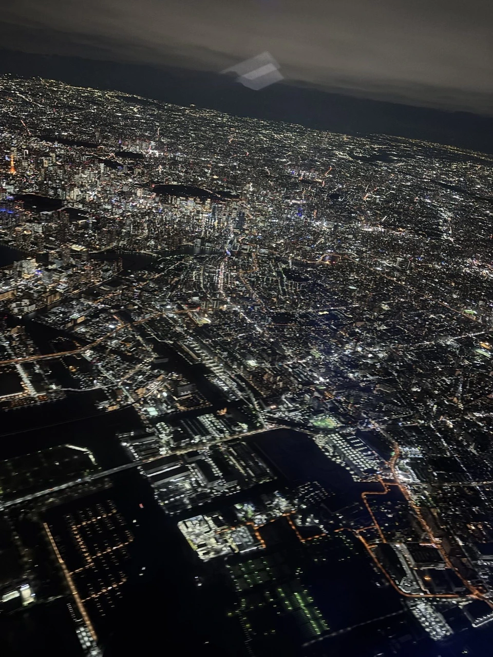 離陸直後の機内の窓から見た東京の夜景。街の明かりが地平線まで広がり、手前に湾岸の埋め立て地