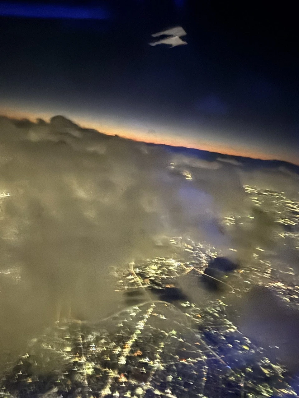 雲の上に出た機内の窓からの景色。雲のすき間に街の明かりが見え、地平線にオレンジ色の夕焼けの線