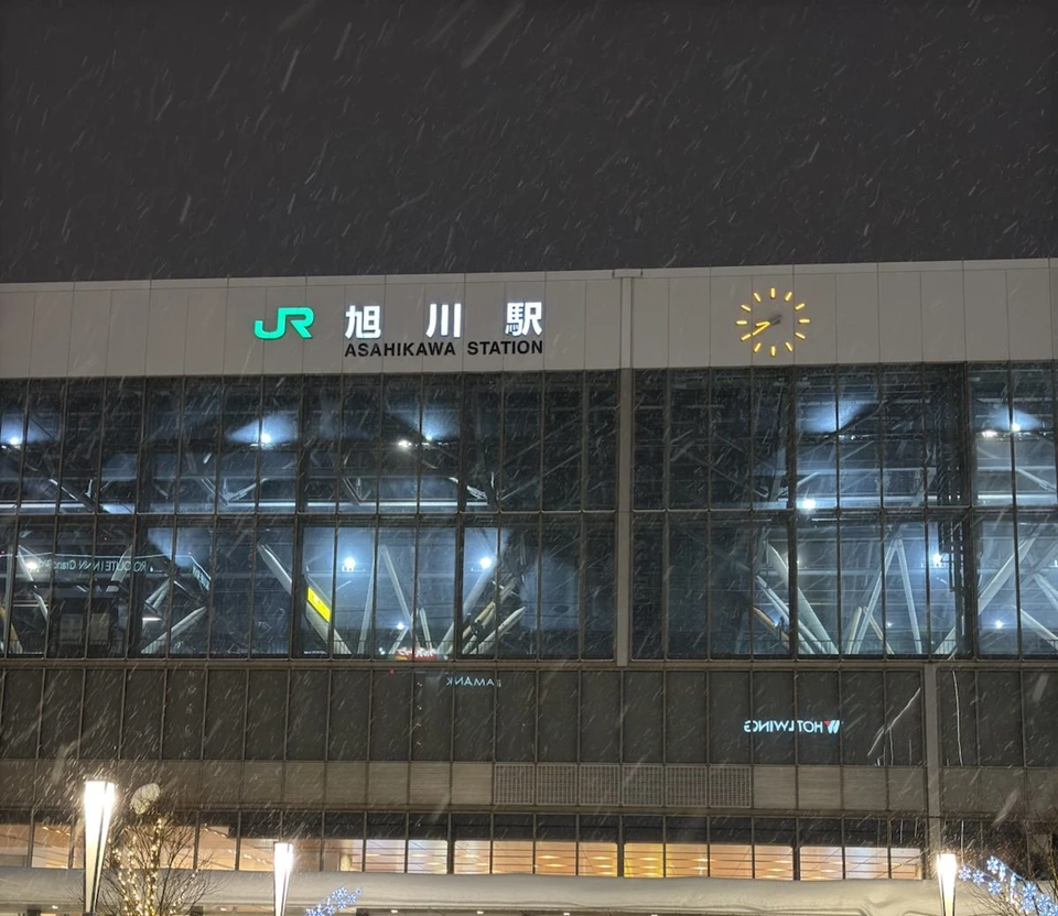 雪の降る夜の旭川駅の駅舎。JR旭川駅の文字と時計、ガラス張りの壁の奥に駅の骨組みが見える