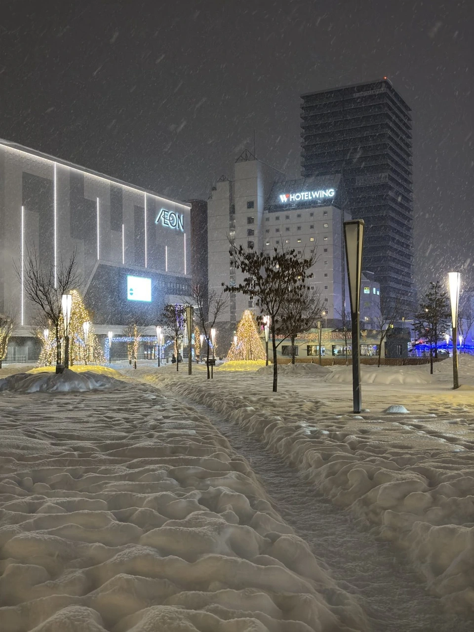 雪の降る夜の旭川駅前広場。雪に足あとの残る広場に金色の電球のツリーが並び、奥にイオンモールとホテルの建物