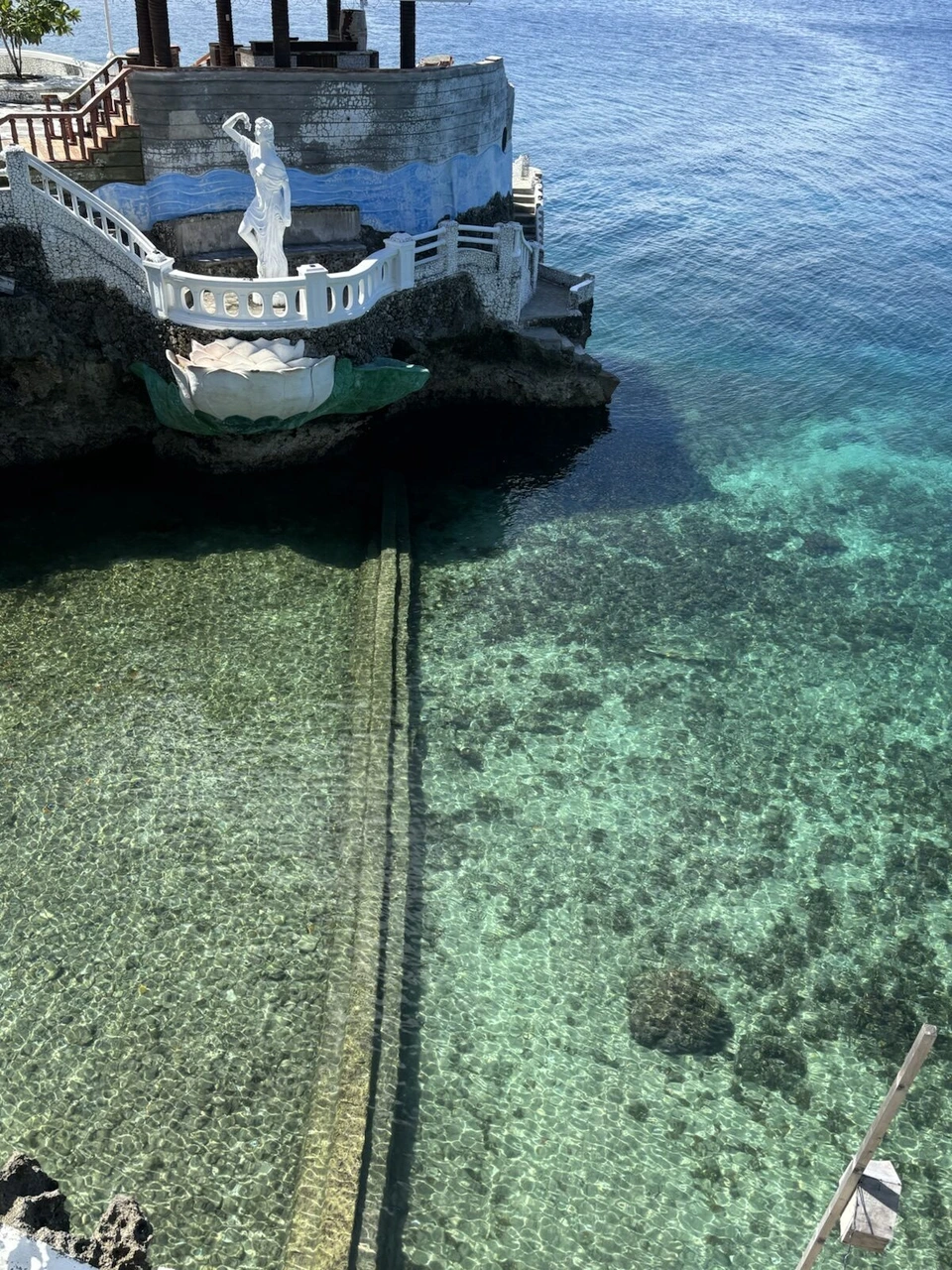崖の上から見下ろした透き通った海と、岩の上の白い像