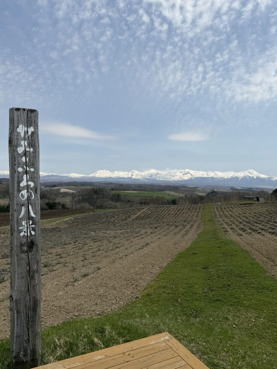 「かみふらの八景」と書かれた木の柱と、畝の並ぶ畑の先の十勝岳連峰