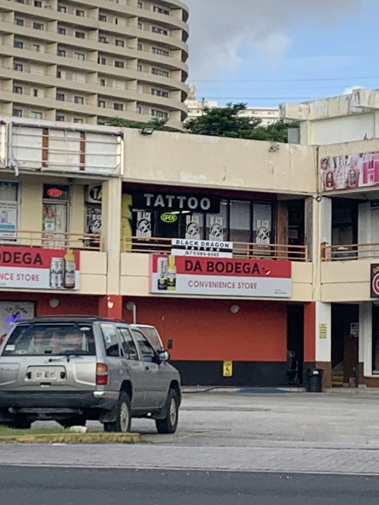 グアム・タモンの通り沿いの2階建ての店舗。タトゥーショップとコンビニの看板、奥に高層ホテル
