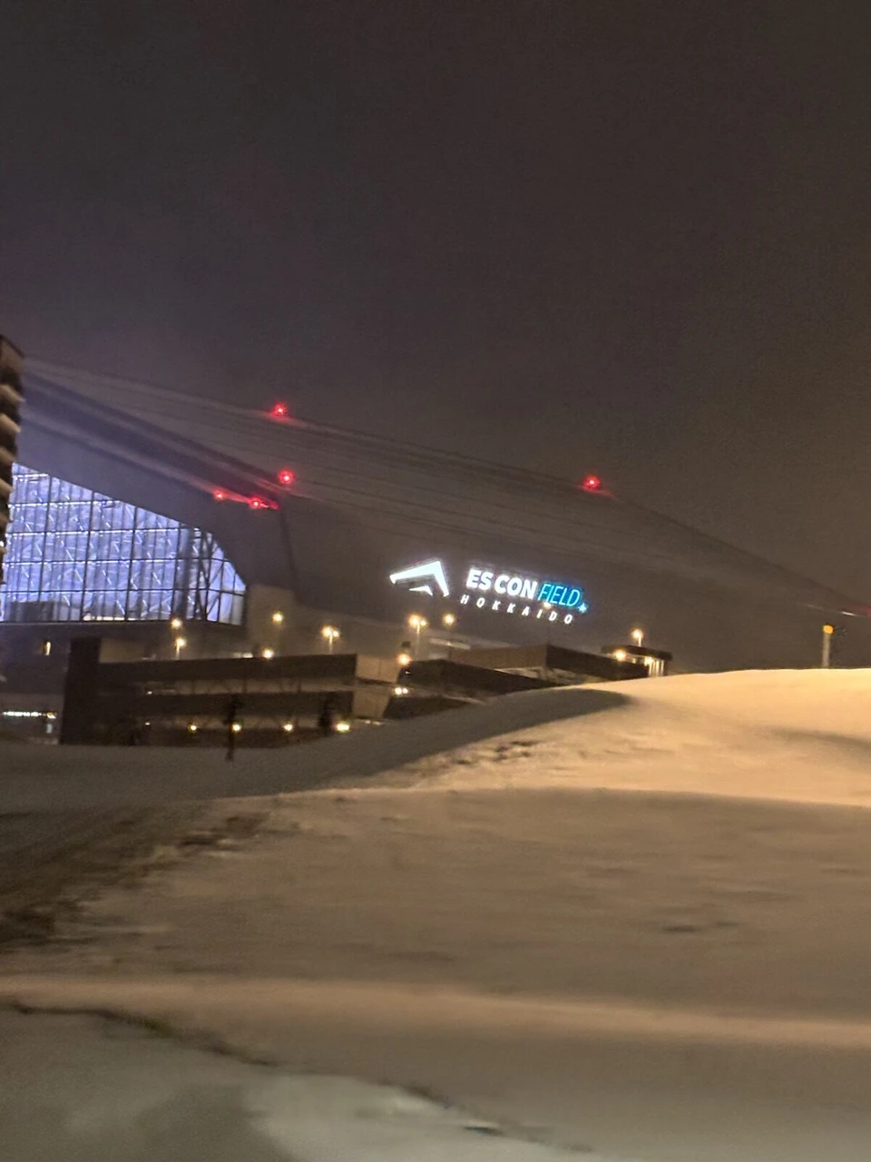雪の丘の向こうに見える夜のエスコンフィールドHOKKAIDO。屋根に赤い航空障害灯