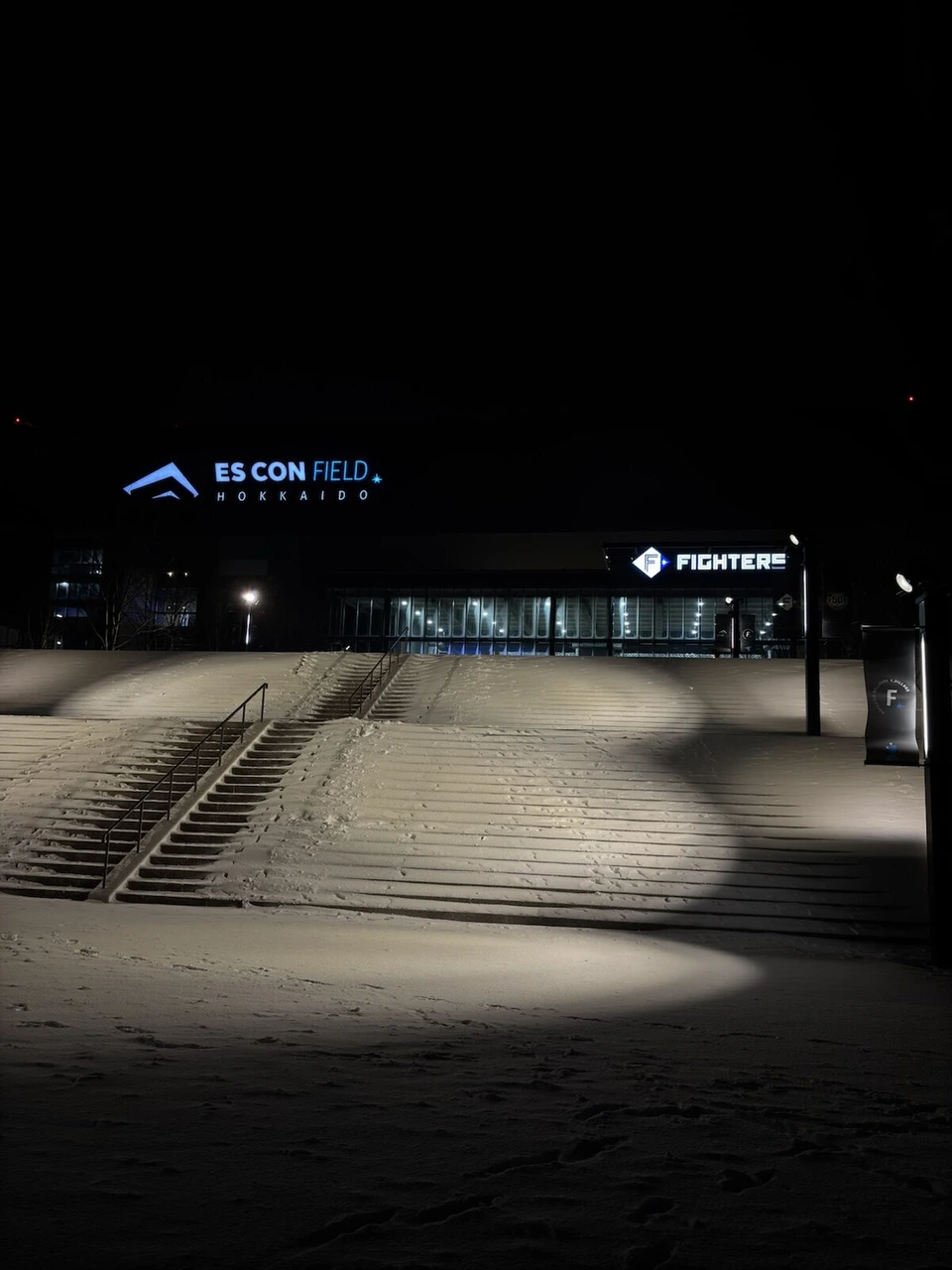 雪に覆われた幅の広い階段と、ライトで丸く照らされた雪面。奥にエスコンフィールドの入口