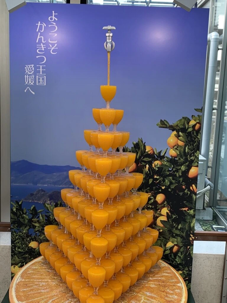 「ようこそかんきつ王国愛媛へ」の看板。グラスを積み上げたみかんジュースのタワー