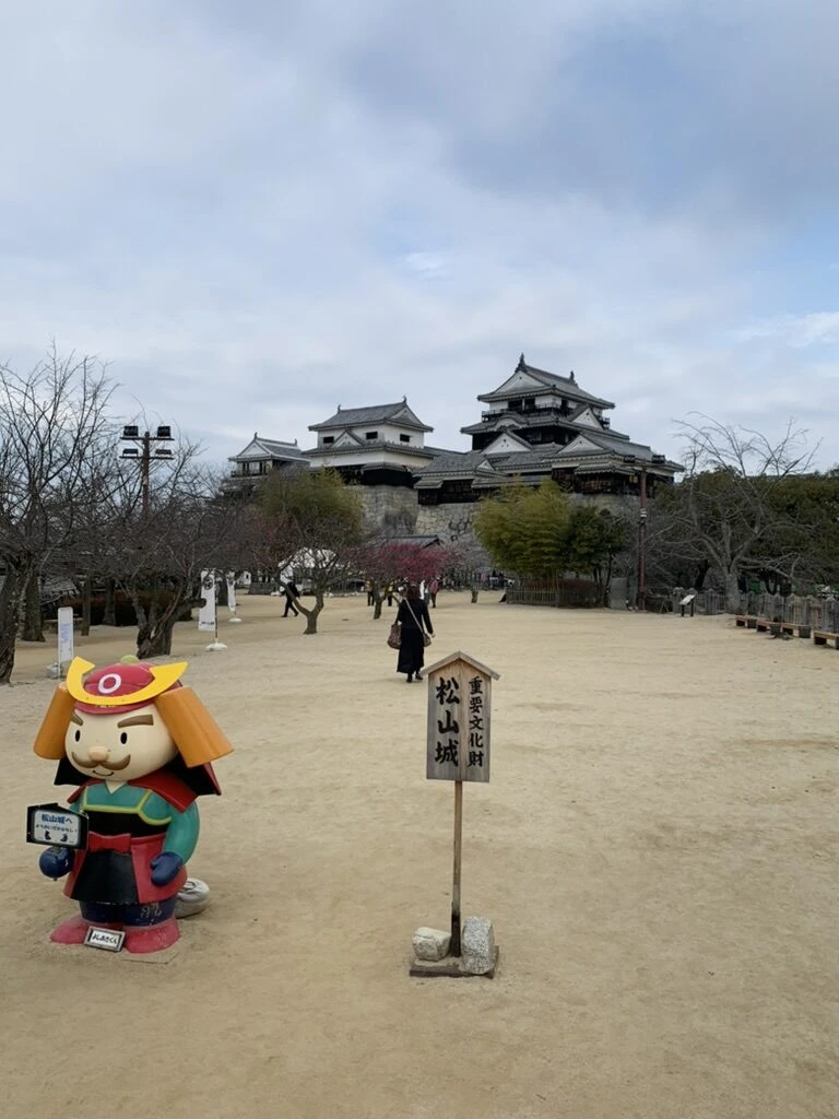 本丸広場の甲冑姿のキャラクター像と「松山城」の標柱。奥に天守