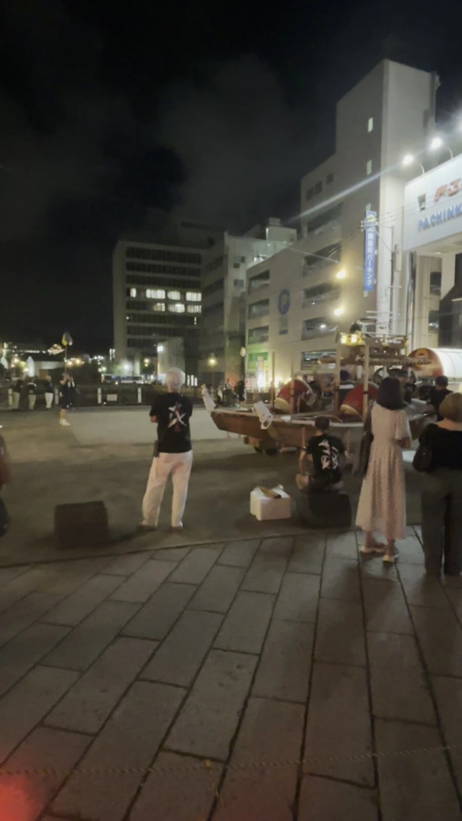 夜の長崎の広場。そろいの黒いTシャツの人たちが船の形の山車を動かしていて、見ている人の後ろ姿